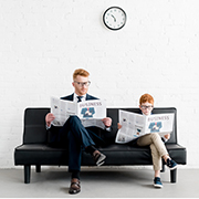 Father and son dressed alike and reading newspaper on couch together
