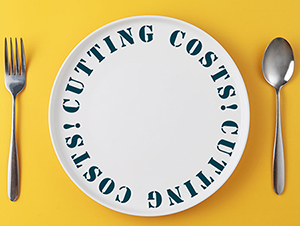 Cutting costs words on a white plate with a fork and spoon on a yellow background SMALL