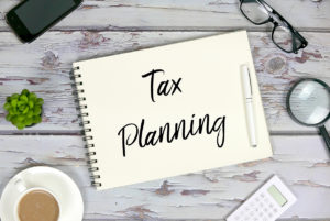 Top view of mobile phone,plant,coffee,calculator,magnifying glass,sunglasses,pen and notebook written with Tax Planning on wooden background.